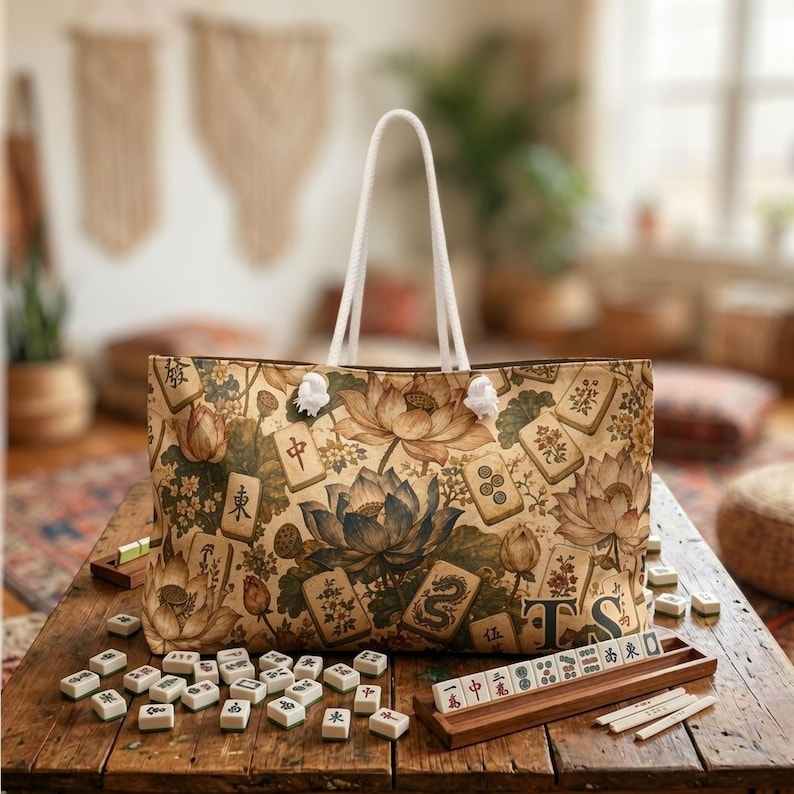May include: A tote bag with white rope handles featuring a beige and brown pattern of lotus flowers and mahjong tiles. The bag displays the letters TS in the bottom right corner. Scattered mahjong tiles and a wooden tile rack are visible.
