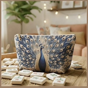 May include: A zipped pouch featuring a blue and cream peacock with a fanned tail, surrounded by floral patterns and a pagoda silhouette, resting among scattered white rectangular tiles with various green and red symbols printed on them.