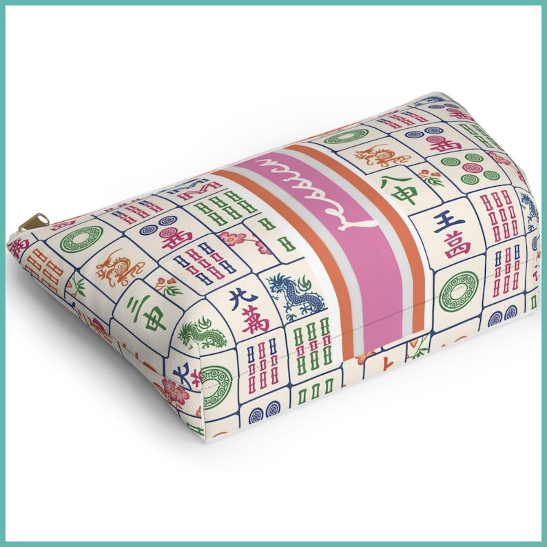 May include: A rectangular cosmetic bag with a zip. The bag has a cream background with a pattern of colourful Mahjong tiles. A pink stripe with the name "Jessica" is centred on the bag.