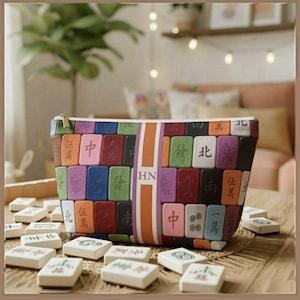 May include: A rectangular zipped pouch featuring a colourful pattern of mahjong tiles in shades of red, blue, green, orange, and purple. A vertical orange and white stripe runs down the centre with the letters HN printed in dark brown.