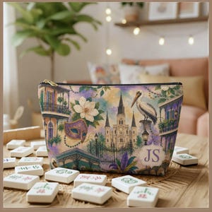 May include: A rectangular zipped pouch featuring a colourful illustration of New Orleans architecture, a pelican, a magnolia flower, and a masquerade mask. The design includes a central label with the monogram JS in a decorative font.