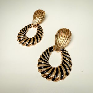 May include: A pair of gold-tone earrings with black enamel detailing. The earrings feature a twisted circle design with a teardrop-shaped pendant.