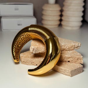 May include: A gold-colored, crescent-shaped bracelet. The bracelet features a textured, honeycomb-like design on one end and a smooth, polished finish on the other. The bracelet is displayed on a stack of beige, rectangular stones.