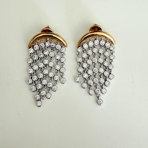 Peut inclure: Une paire de boucles d'oreilles élégantes avec un dessus en croissant doré et des chaînes argentées en cascade. Chaque chaîne comporte de multiples pierres précieuses rondes et claires, créant un effet de lustre étincelant. Boucles d'oreilles percées.