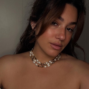 May include: A woman wearing a white top with a gold and pearl necklace. The necklace has two strands of pearls and a gold chain.