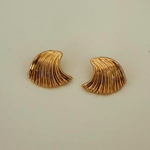 May include: A pair of gold-coloured seashell-shaped stud earrings. The earrings have a textured, ridged surface, and a curved, fan-like design. The earrings are set against a neutral background.