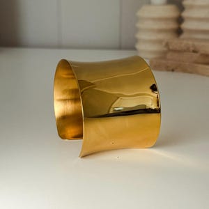 May include: A wide, gold-colored cuff bracelet with a curved design. The polished metal reflects light, creating highlights and shadows. The bracelet is a statement piece, suitable for adding a touch of elegance to any outfit.