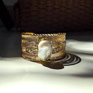 May include: A wide gold cuff bracelet with an oval, off-white pearl centerpiece. The bracelet has a textured, woven design with intricate patterns. The cuff sits on a white surface, with shadows cast across it.