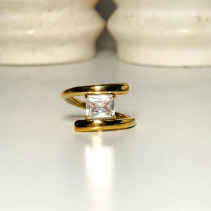 May include: A gold ring with a square-shaped clear stone set in the center. The ring has a unique twisted design.