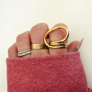 May include: A hand wearing a set of gold rings. The rings are stacked on the middle finger and include a thin band with small diamonds, a thicker band with a small circle design, and a large, open, twisted ring.
