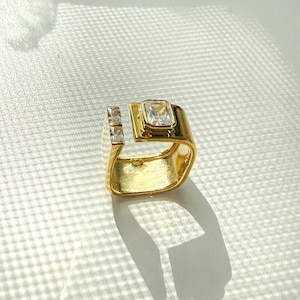 May include: A gold-colored ring with a split shank design. The ring features a large, square, clear gemstone and two smaller, rectangular gemstones. The ring is set on a white surface.