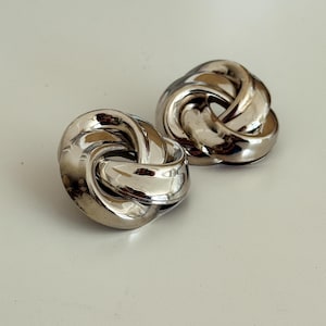 Peut inclure: Une paire de boucles d'oreilles en argent en forme de nœud. Les boucles d'oreilles sont en métal lisse et brillant et ont un design simple et élégant.