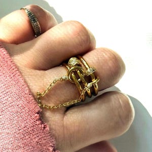 Peut inclure: Une main portant plusieurs bagues dorées. Une bague est recouverte de petites pierres scintillantes. Une autre bague présente un motif de nœud, relié par une chaîne à une bague sur le doigt adjacent. Les bagues sont de couleur or vif.