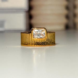 May include: A gold ring with a clear rectangular stone set in a raised frame. The band of the ring has a ridged texture.
