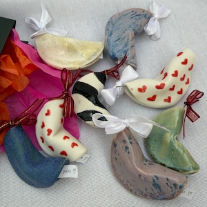 May include: A collection of ceramic fortune cookie ornaments in various colours and patterns, including red hearts, black stripes, and solid blue, green, and speckled glazes. Each cookie is tied with a small white or red ribbon and has a paper tag.