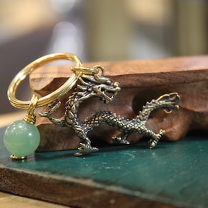 Puede incluir: Un llavero dorado con una cuenta de jade verde y un amuleto de dragón detallado.
