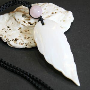May include: A white pendant necklace with a leaf-shaped design, strung on a black cord. A small, pink, round bead accents the top of the pendant. The necklace rests on a white, textured seashell against a dark background.
