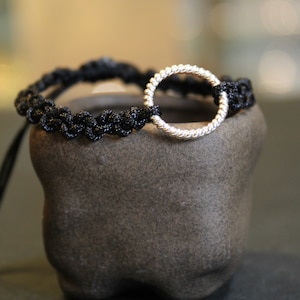 May include: A black braided bracelet with a silver ring charm. The bracelet is on a dark gray textured surface.