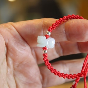 May include: Red macrame bracelet featuring a small, light green jade carving of an animal. The bracelet has silver-colored beads and a sliding knot closure. The bracelet is held in a hand.
