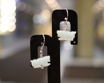 May include: A pair of silver earrings with white jade pig charms. The earrings are hanging from a black display stand.