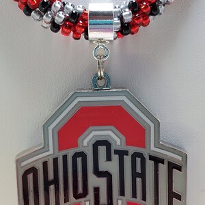 May include: A red, white, and gray Ohio State University logo charm necklace with a silver toggle clasp. The necklace is made of black, red, and silver beads.