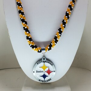 Pode incluir: Um colar de contas pretas, brancas e douradas com um pingente do Pittsburgh Steelers. O pingente &eacute; de prata com o logotipo da equipe e as palavras "Pittsburgh Steelers".