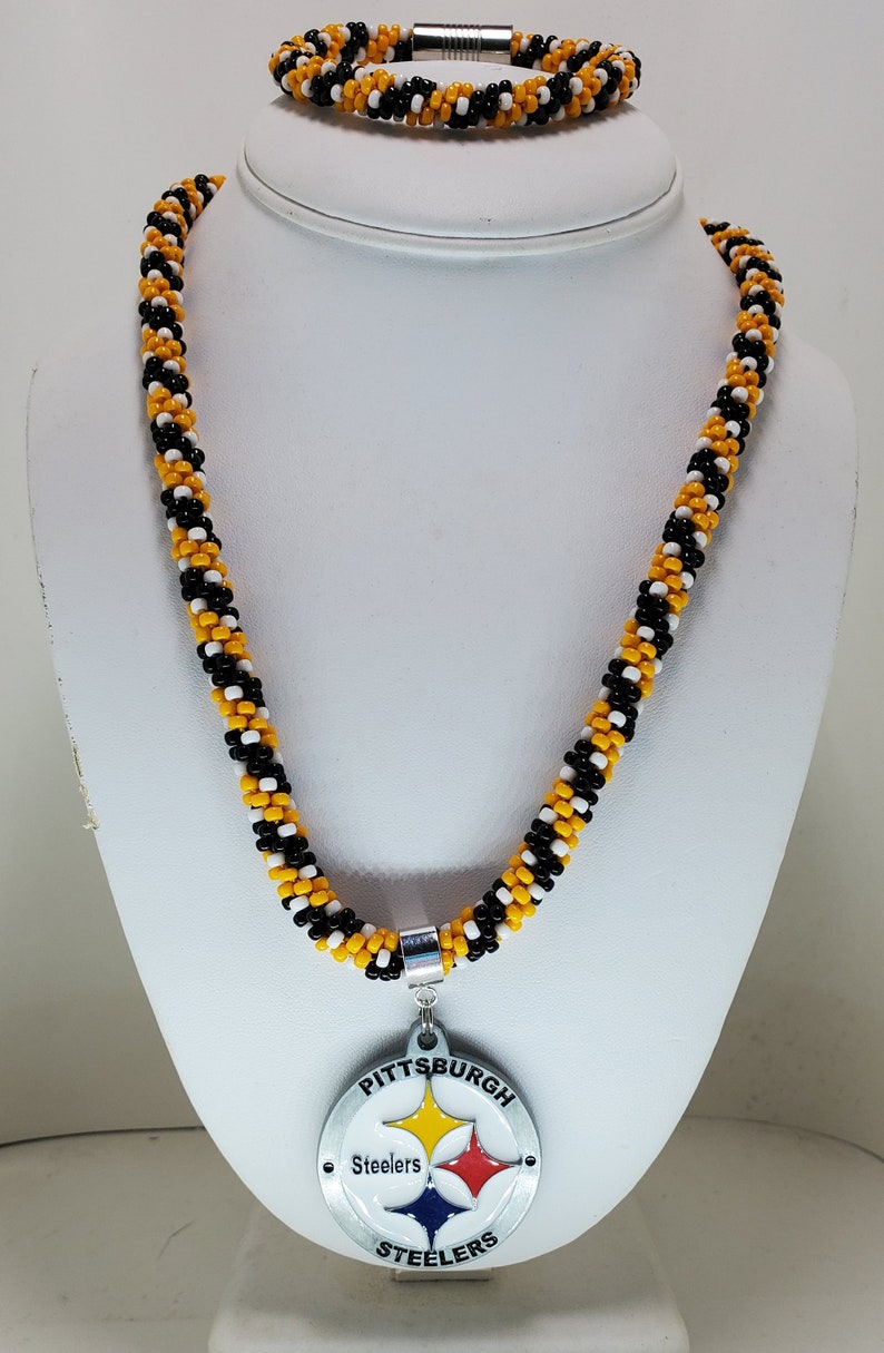 Pode incluir: Um colar de contas pretas, brancas e douradas com um pingente do Pittsburgh Steelers. O pingente &eacute; de prata com o logotipo da equipe e as palavras "Pittsburgh Steelers".