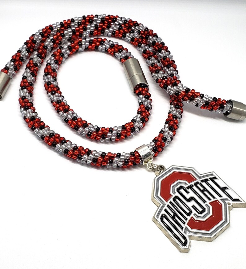 May include: A red, black, and silver beaded necklace with a silver magnetic clasp and a silver charm with the Ohio State logo. The charm is red, white, and gray with the words "Ohio State" in black.