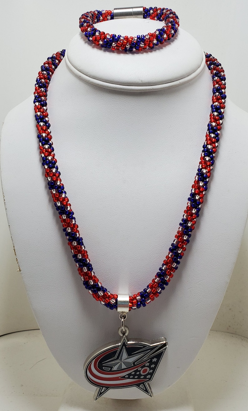 May include: A red, white, and blue beaded necklace and bracelet set with a silver charm featuring the Columbus Blue Jackets NHL team logo.