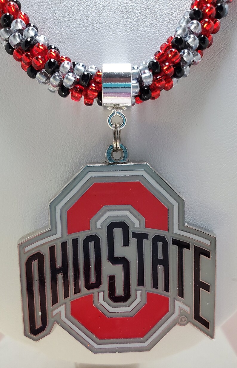 May include: A red, white, and gray Ohio State University logo charm necklace with a silver toggle clasp. The necklace is made of black, red, and silver beads.