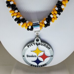 Pode incluir: Um colar de contas pretas e amarelas com um pingente prateado do Pittsburgh Steelers. O pingente apresenta o logotipo da equipe e as palavras "Pittsburgh Steelers".