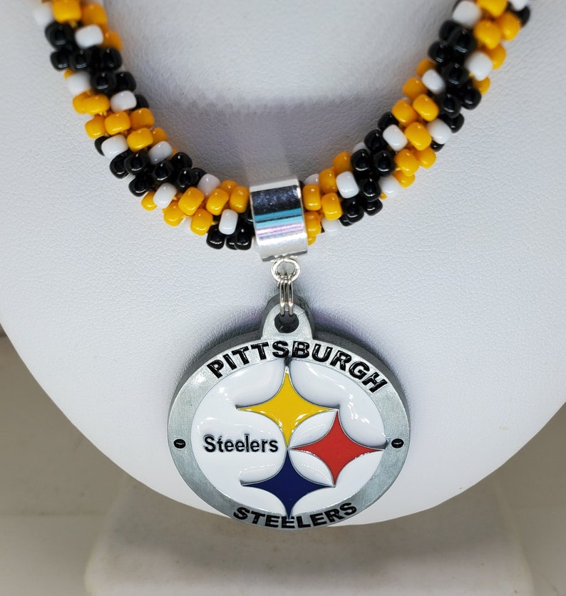 Pode incluir: Um colar de contas pretas e amarelas com um pingente prateado do Pittsburgh Steelers. O pingente apresenta o logotipo da equipe e as palavras "Pittsburgh Steelers".