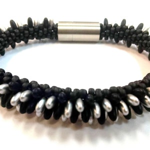 May include: A black and silver beaded bracelet with a magnetic clasp. The bracelet is made with small, round beads and has a unique, textured design.