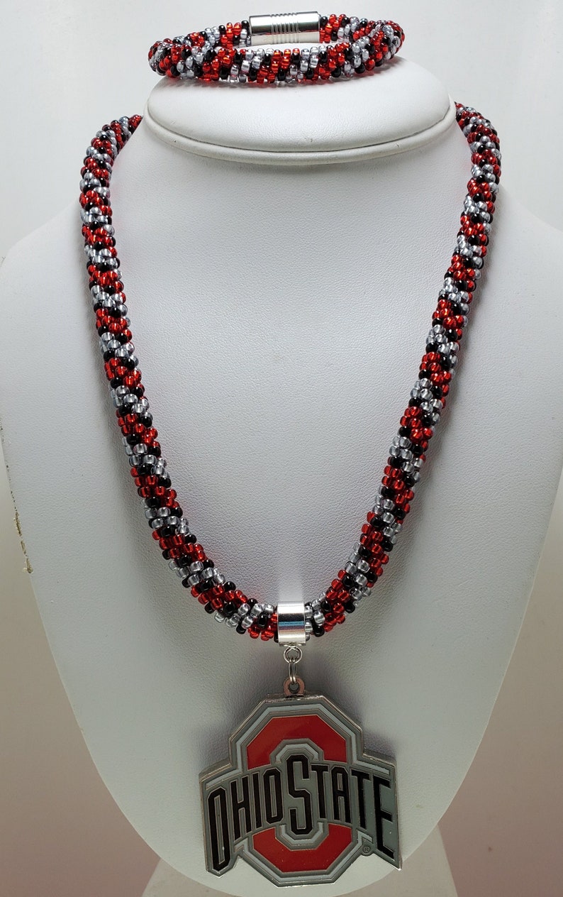 May include: A red, black, and silver beaded necklace and bracelet set with a silver charm featuring the Ohio State logo.