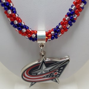May include: A red, white, and blue beaded necklace with a silver charm featuring the Columbus Blue Jackets NHL team logo. The logo is a silver star with a red, white, and blue design.