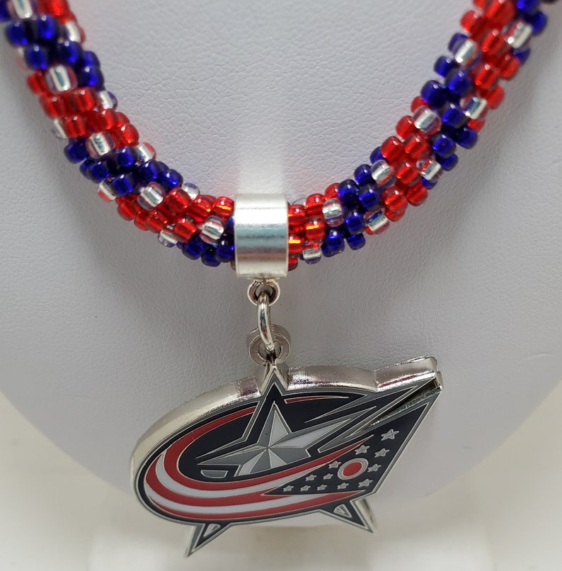 May include: A red, white, and blue beaded necklace with a silver charm featuring the Columbus Blue Jackets NHL team logo. The logo is a silver star with a red, white, and blue design.