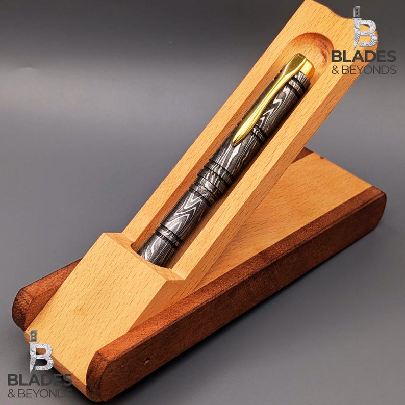 Puede incluir: Un bol&iacute;grafo artesanal con un cuerpo de metal estampado y detalles dorados descansa en un estuche de madera. El estuche est&aacute; hecho de madera clara y oscura, con el texto "Blades & Beyonds" visible.