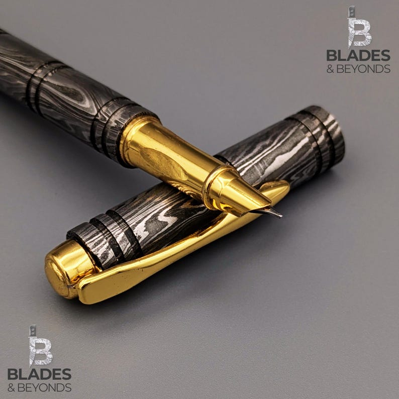 Puede incluir: Un bol&iacute;grafo roller de acero de Damasco con clip y detalles dorados. El cuerpo del bol&iacute;grafo presenta un patr&oacute;n &uacute;nico de remolinos oscuros y claros. La tapa del bol&iacute;grafo est&aacute; colocada junto al bol&iacute;grafo, revelando la punta. El logotipo "Blades & Beyonds" es visible.