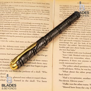 Puede incluir: Una pluma estilogr&aacute;fica de lujo con un patr&oacute;n veteado negro y gris, detalles dorados y un agarre texturizado. La pluma se muestra sobre un libro abierto con texto impreso. La pluma es un instrumento de escritura.