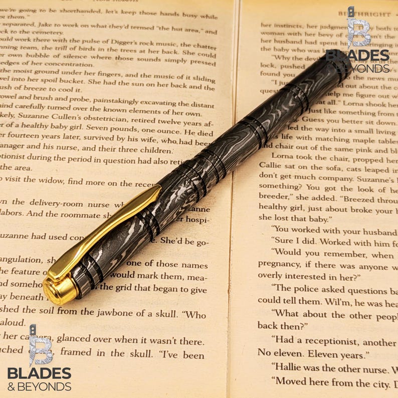 Puede incluir: Una pluma estilogr&aacute;fica de lujo con un patr&oacute;n veteado negro y gris, detalles dorados y un agarre texturizado. La pluma se muestra sobre un libro abierto con texto impreso. La pluma es un instrumento de escritura.