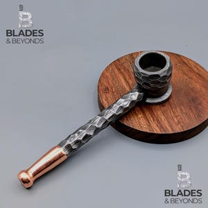 May include: A handcrafted pipe with a textured, dark gray finish and a copper-colored mouthpiece. The pipe rests on a round, wooden base. The image includes the text "BLADES & BEYONDS".