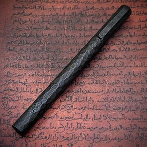 May include: A black fountain pen with a textured, hammered metal finish. The pen has a hexagonal barrel and a cap. The background is a reddish-brown surface with faded text.