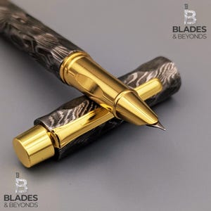 Op de afbeelding: Een luxe rollerball pen met een patroon, donkergrijze en zwarte barrel en een goudkleurige metalen dop en accenten. De pen ligt op zijn kant, met de dop gedeeltelijk verwijderd. De pen heeft de tekst "BLADES & BEYONDS".