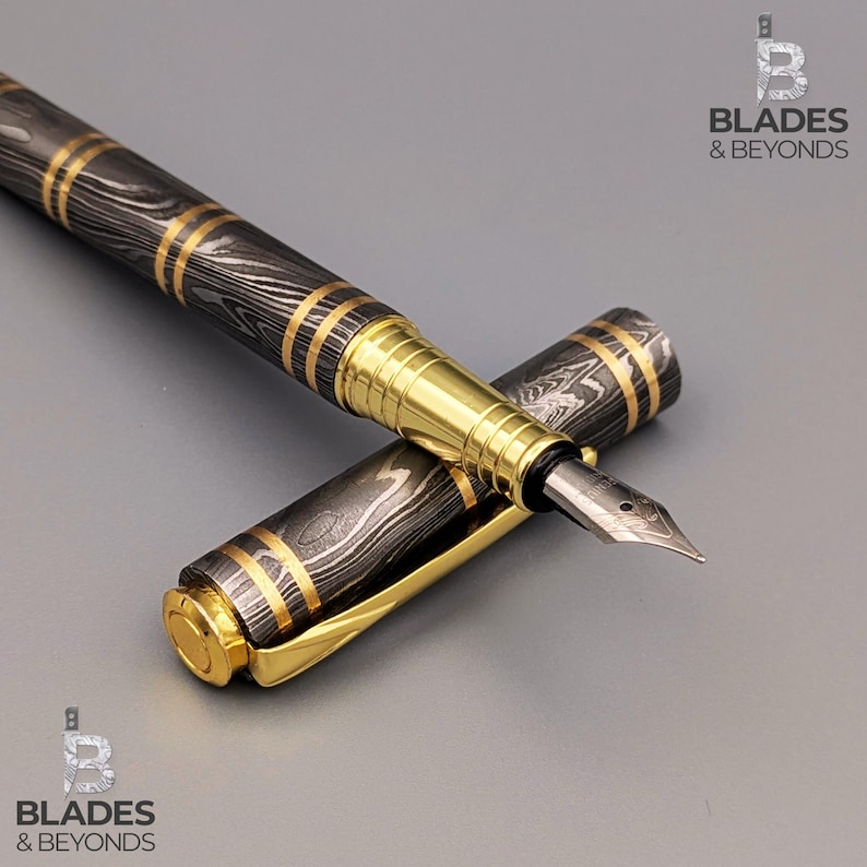 May include: A luxury fountain pen with a black and silver patterned barrel and gold accents. The pen's cap is off, revealing the nib. The pen is lying on a gray surface. The pen has the text "Blades & Beyonds" on the image.