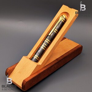 May include: A black and gold pen with a wood grain pattern rests in a wooden pen case. The pen has gold accents and is displayed in a light brown wooden case with a dark brown base. The pen case is open, showcasing the pen.