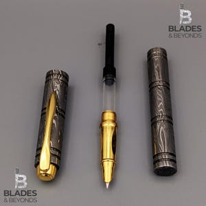 Puede incluir: Un bol&iacute;grafo desmontado con plum&iacute;n y detalles dorados, un dep&oacute;sito de tinta transparente y un cuerpo y capuch&oacute;n de metal estampado. El capuch&oacute;n del bol&iacute;grafo tiene un clip dorado. El bol&iacute;grafo se muestra sobre una superficie gris. El logotipo "Blades & Beyonds" es visible.