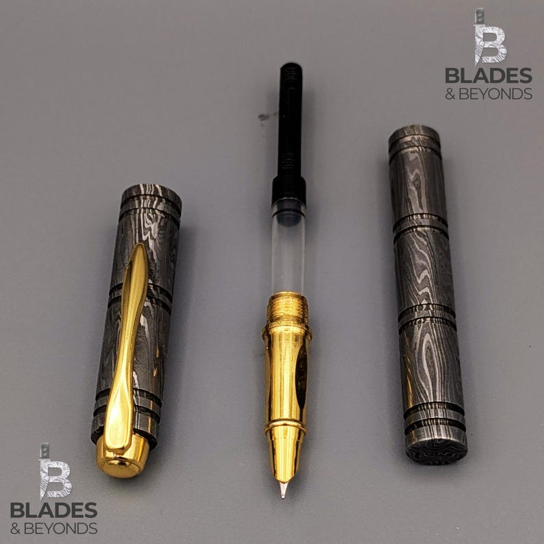 Puede incluir: Un bol&iacute;grafo desmontado con plum&iacute;n y detalles dorados, un dep&oacute;sito de tinta transparente y un cuerpo y capuch&oacute;n de metal estampado. El capuch&oacute;n del bol&iacute;grafo tiene un clip dorado. El bol&iacute;grafo se muestra sobre una superficie gris. El logotipo "Blades & Beyonds" es visible.