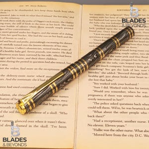May include: A luxury fountain pen with a black and silver patterned barrel and gold-colored accents. The pen is lying on an open book with text. The pen is a writing instrument.