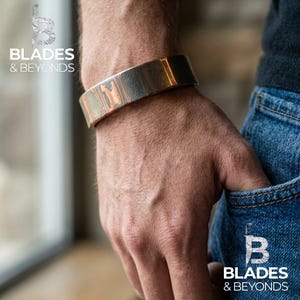 Puede incluir: Un brazalete metálico ancho y estampado, con tonos cobrizos y plateados. El brazalete se lleva en la muñeca. El texto "BLADES & BEYONDS" es visible en la imagen.