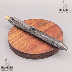 Puede incluir: Un bolígrafo de acero de Damasco con punta de latón y clip plateado descansa sobre una superficie de madera redonda. El bolígrafo tiene un patrón único y está inclinado hacia la derecha. Es un instrumento de escritura.